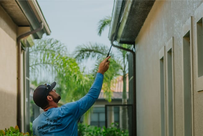 Technician from Elegant Styles Power Wash removing moss and lichen from soffits on a Naples, Florida home using professional equipment.