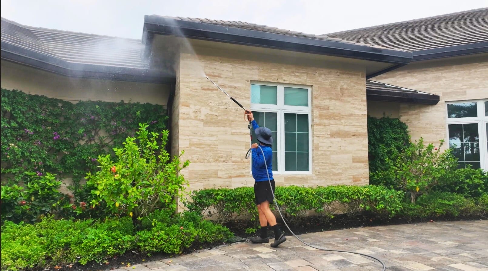 Technician wearing safety gear cleaning debris from a white soffit and gutter under a Southwest Florida home’s roofline using a power washing wand.